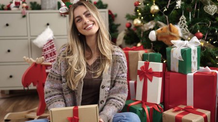 Photo for Young blonde woman holding gift sitting by christmas tree at home - Royalty Free Image