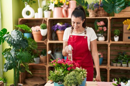 Téléchargez les photos : Jeune belle hispanique femme fleuriste en utilisant diffuseur arrosage plante à la boutique de fleurs - en image libre de droit