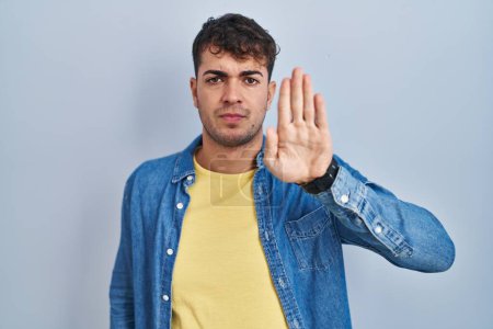 Photo for Young hispanic man standing over blue background doing stop sing with palm of the hand. warning expression with negative and serious gesture on the face. - Royalty Free Image