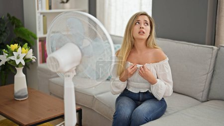Téléchargez les photos : Jeune femme blonde assise sur un canapé souffrant de chaleur en utilisant un ventilateur à la maison - en image libre de droit