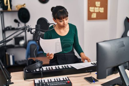 Téléchargez les photos : Jeune belle hispanique femme musicien lecture document assis sur chaise au studio de musique - en image libre de droit
