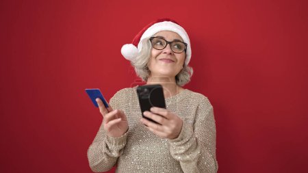 Photo for Middle age woman with grey hair doing christmas online shopping with smartphone over isolated red background - Royalty Free Image