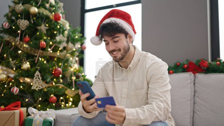 Photo for Young hispanic man shopping with smartphone and credit card celebrating christmas at home - Royalty Free Image