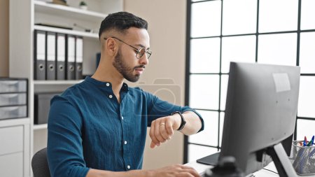 Téléchargez les photos : Jeune homme hispanique travailleur d'affaires en utilisant l'ordinateur en regardant l'heure au bureau - en image libre de droit