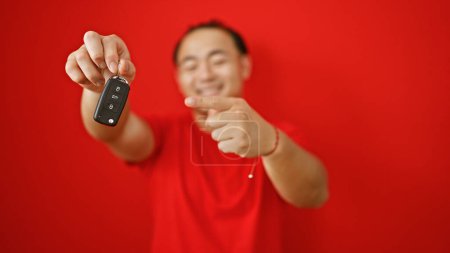 Téléchargez les photos : Heureux jeune chinois pointant avec enthousiasme les clés de sa nouvelle voiture, heureusement isolé sur fond rouge - en image libre de droit