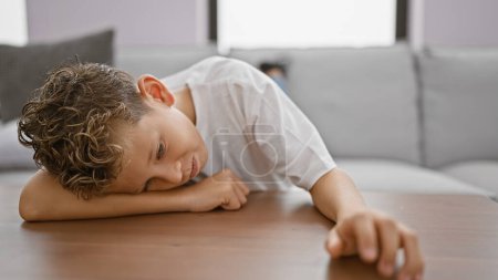 Photo for Adorable little blond boy, with a sad expression, comfortably leaning on a living room table. a pensive, serious indoor atmosphere looms within the cozy home. - Royalty Free Image