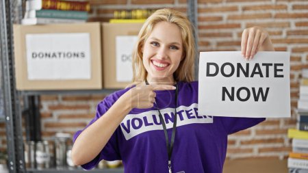 Téléchargez les photos : Jeune femme blonde bénévole souriant confiant pointant pour faire un don maintenant bannière au centre de bienfaisance - en image libre de droit