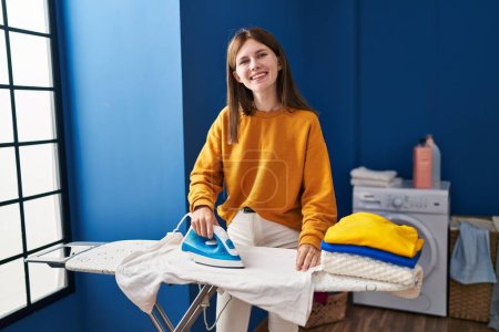 Foto de Mujer rubia joven sonriendo confiado planchar ropa en la sala de lavandería - Imagen libre de derechos