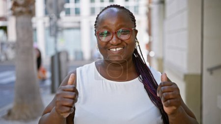 Photo for Joyful, confident african american woman flashing a fun, approving thumbs-up sign on a sunny city street, her cool glasses gleaming. - Royalty Free Image