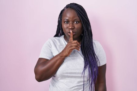 Téléchargez les photos : Jeune femme africaine debout sur fond rose demandant à être tranquille avec le doigt sur les lèvres. silence et concept secret. - en image libre de droit