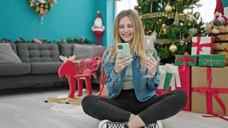 Photo for Young blonde woman celebrating christmas holding champagne using smartphone at home - Royalty Free Image
