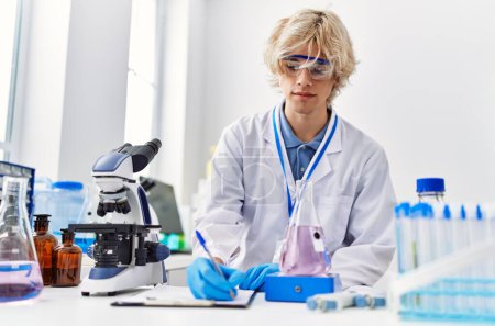 Téléchargez les photos : Jeune homme blond scientifique écrit sur un document pesant une éprouvette au laboratoire - en image libre de droit