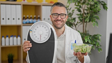 Foto de Hombre de pelo gris dietista sosteniendo ensalada y pesadora en la clínica - Imagen libre de derechos