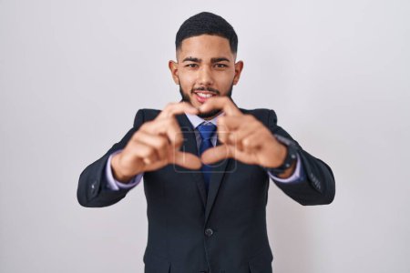 Foto de Joven hombre hispano vistiendo traje de negocios y corbata sonriendo en amor haciendo forma de símbolo del corazón con las manos. concepto romántico. - Imagen libre de derechos