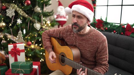 Foto de Joven calvo tocando la guitarra clásica sentado en el sofá por el árbol de Navidad en casa - Imagen libre de derechos
