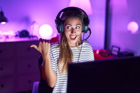 Téléchargez les photos : Jeune femme blonde jouant à des jeux vidéo pointant le pouce vers le côté souriant heureux avec la bouche ouverte - en image libre de droit