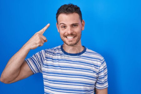 Foto de Young hispanic man standing over blue background smiling pointing to head with one finger, great idea or thought, good memory - Imagen libre de derechos