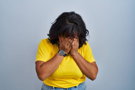 Foto de Mujer hispana de pie sobre fondo azul con expresión triste cubriendo la cara con las manos mientras llora. concepto de depresión. - Imagen libre de derechos