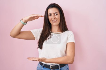 Foto de Mujer morena joven de pie sobre fondo rosa gesto con las manos mostrando signo de tamaño grande y grande, símbolo de medida. sonriendo mirando a la cámara. concepto de medición. - Imagen libre de derechos