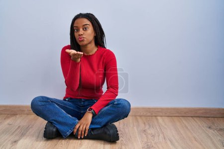 Téléchargez les photos : Jeune afro-américain avec des tresses assis sur le sol à la maison en regardant la caméra souffler un baiser avec la main sur l'air étant belle et sexy. expression d'amour. - en image libre de droit