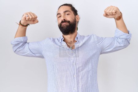 Téléchargez les photos : Homme hispanique avec barbe portant une chemise décontractée montrant les muscles des bras souriant fier. concept de fitness. - en image libre de droit