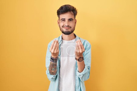 Téléchargez les photos : Jeune homme hispanique avec des tatouages debout sur fond jaune faisant geste d'argent avec les mains, demandant le paiement du salaire, entreprise millionnaire - en image libre de droit