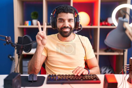 Téléchargez les photos : Homme hispanique avec barbe jouant à des jeux vidéo avec écouteurs souriant avec un sourire heureux clin d'oeil à la caméra faisant signe de victoire. numéro deux. - en image libre de droit