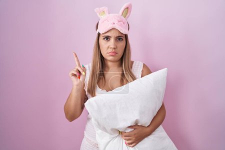 Foto de Mujer rubia joven con pijama abrazando almohada apuntando hacia arriba buscando triste y molesto, indicando la dirección con los dedos, infeliz y deprimido. - Imagen libre de derechos