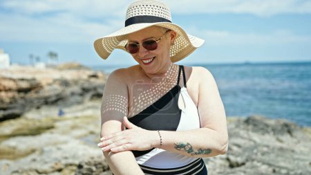 Téléchargez les photos : Femme aux cheveux gris d'âge moyen portant un maillot de bain et un chapeau d'été appliquant de la crème solaire sur le bras à la plage - en image libre de droit