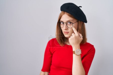 Téléchargez les photos : Jeune rousse femme debout portant des lunettes et un béret pointant vers l'œil vous regardant geste, expression suspecte - en image libre de droit