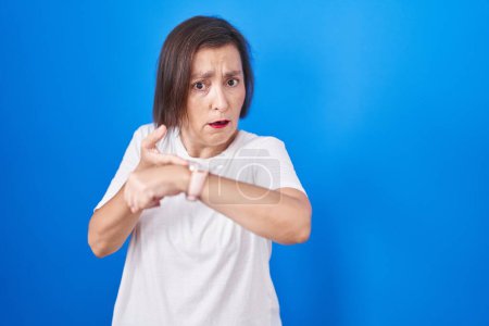 Téléchargez les photos : Middle age hispanic woman standing over blue background in hurry pointing to watch time, impatience, upset and angry for deadline delay - en image libre de droit
