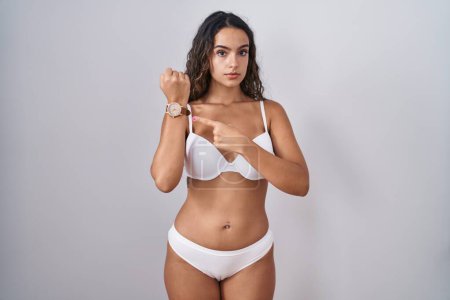Téléchargez les photos : Jeune femme hispanique portant de la lingerie blanche à la hâte pointant vers regarder le temps, l'impatience, regardant la caméra avec une expression détendue - en image libre de droit