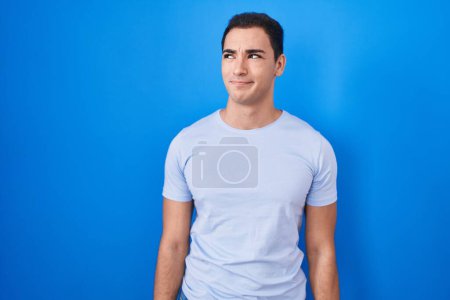Foto de Joven hombre hispano de pie sobre fondo azul sonriendo mirando al costado y mirando hacia otro lado pensando. - Imagen libre de derechos