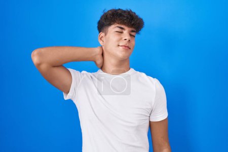 Téléchargez les photos : Adolescent hispanique debout sur fond bleu souffrant d'une blessure au cou, toucher le cou avec la main, douleur musculaire - en image libre de droit
