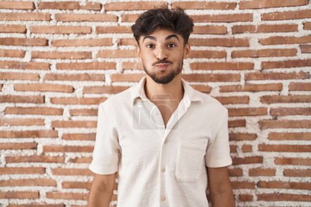 Photo for Arab man with beard standing over bricks wall background puffing cheeks with funny face. mouth inflated with air, crazy expression. - Royalty Free Image