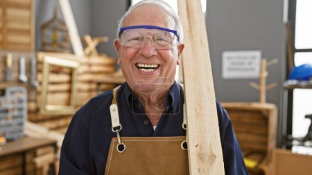 Photo for Confident senior man, a professional carpenter, smiling while securely holding a wood plank in carpentry workshop - Royalty Free Image