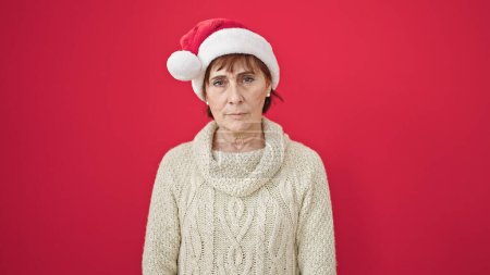 Photo for Mature hispanic woman standing with serious expression wearing christmas hat over isolated red background - Royalty Free Image