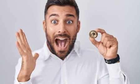 Photo for Handsome hispanic man holding polkadot cryptocurrency coin celebrating victory with happy smile and winner expression with raised hands - Royalty Free Image
