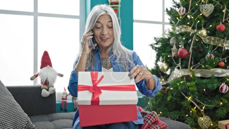 Foto de Mujer de pelo gris de mediana edad desempacando regalo de Navidad hablando en el teléfono inteligente en casa - Imagen libre de derechos