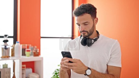 Photo for Young hispanic man using smartphone wearing headphones standing at dinning room - Royalty Free Image