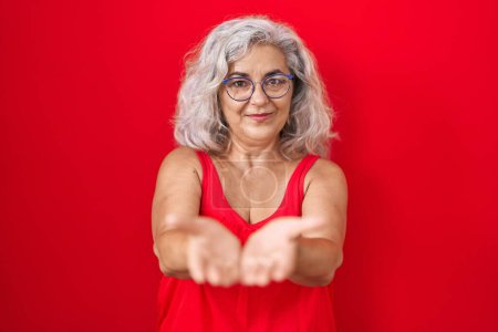 Foto de Mujer de mediana edad con el pelo gris de pie sobre fondo rojo sonriendo con las palmas de las manos juntas recibiendo o dando gesto. retención y protección - Imagen libre de derechos