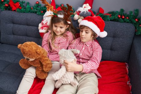 Photo for Brother and sister playing with teddy bears sitting on sofa by christmas decoration at home - Royalty Free Image