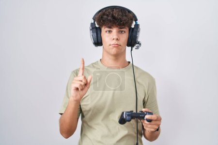 Téléchargez les photos : Adolescent hispanique jouant le contrôleur de tenue de jeu vidéo pointant vers le haut regardant triste et contrarié, indiquant la direction avec les doigts, malheureux et déprimé. - en image libre de droit