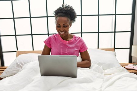 Photo for African american woman using laptop sitting on bed at bedroom - Royalty Free Image