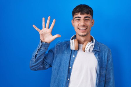 Foto de Joven hombre hispano de pie sobre fondo azul mostrando y señalando hacia arriba con los dedos número cinco mientras sonríe confiado y feliz. - Imagen libre de derechos