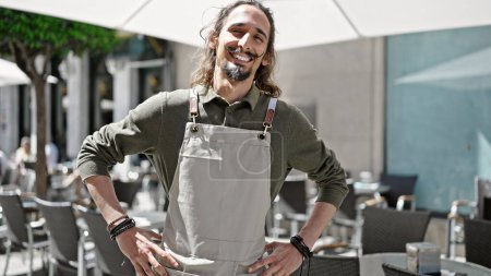 Foto de Joven hombre hispano camarero sonriendo confiado de pie en la terraza de la cafetería - Imagen libre de derechos