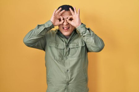 Foto de Joven hispano de pie sobre fondo amarillo haciendo un buen gesto como binoculares sacando la lengua, ojos mirando a través de los dedos. expresión loca. - Imagen libre de derechos