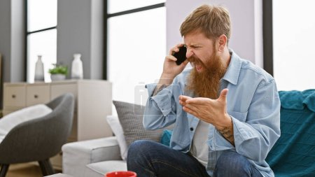 Photo for Unhappy young redhead man, argument escalating while using phone on sofa, expression shows disagreement, home setting amplifying problem, adds serious tone to indoor conversation - Royalty Free Image