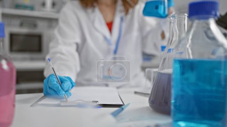 Téléchargez les photos : Jeune femme scientifique concentrée analysant le liquide dans une éprouvette, prenant des notes dans un laboratoire moderne efficace - en image libre de droit