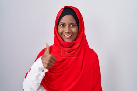 Téléchargez les photos : Jeune femme arabe portant une écharpe traditionnelle islamique en hijab faisant des pouces heureux geste avec la main. approuvant l'expression regardant la caméra montrant le succès. - en image libre de droit